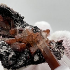 Puede incluir: Primer plano de un esp&eacute;cimen mineral con grupos de cristales de color marr&oacute;n rojizo. Los cristales est&aacute;n bien definidos y tienen un aspecto v&iacute;treo, sobre una matriz oscura y texturizada. La muestra est&aacute; bien iluminada, destacando la estructura del cristal.