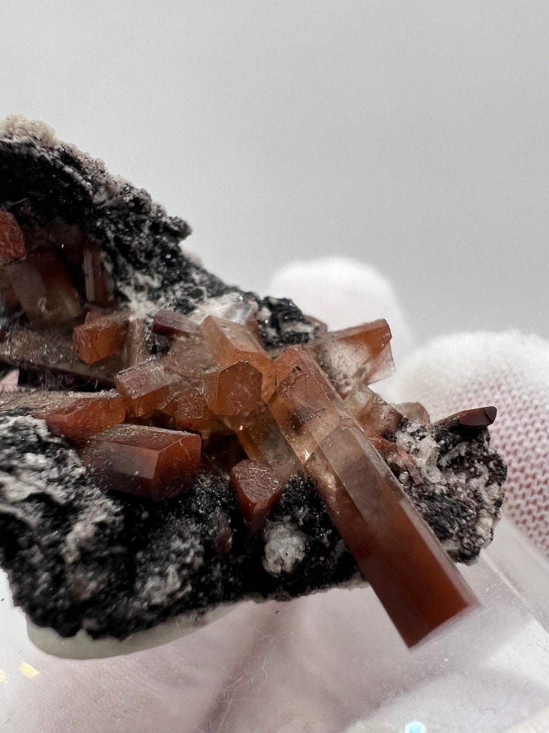 Puede incluir: Primer plano de un esp&eacute;cimen mineral con grupos de cristales de color marr&oacute;n rojizo. Los cristales est&aacute;n bien definidos y tienen un aspecto v&iacute;treo, sobre una matriz oscura y texturizada. La muestra est&aacute; bien iluminada, destacando la estructura del cristal.