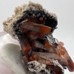 Puede incluir: Primer plano de un esp&eacute;cimen mineral con grupos de cristales de color marr&oacute;n rojizo. Los cristales se encuentran sobre una matriz negra y blanca. La formaci&oacute;n mineral est&aacute; sobre una superficie blanca.