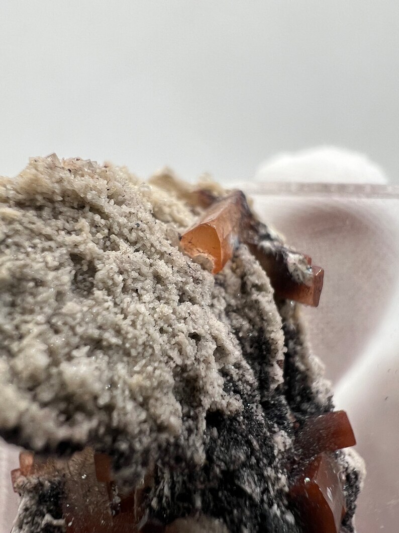 Puede incluir: Primer plano de un esp&eacute;cimen mineral con peque&ntilde;os cristales transl&uacute;cidos de color marr&oacute;n anaranjado. Los cristales se encuentran sobre un fondo de roca rugosa, granular, gris claro y negra. La textura general es &aacute;spera y detallada.