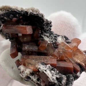 Puede incluir: Primer plano de un esp&eacute;cimen mineral con cristales de color marr&oacute;n rojizo, de forma rectangular. Estos cristales est&aacute;n colocados sobre una base oscura y texturizada con un revestimiento blanco y granulado. Los cristales son transl&uacute;cidos.