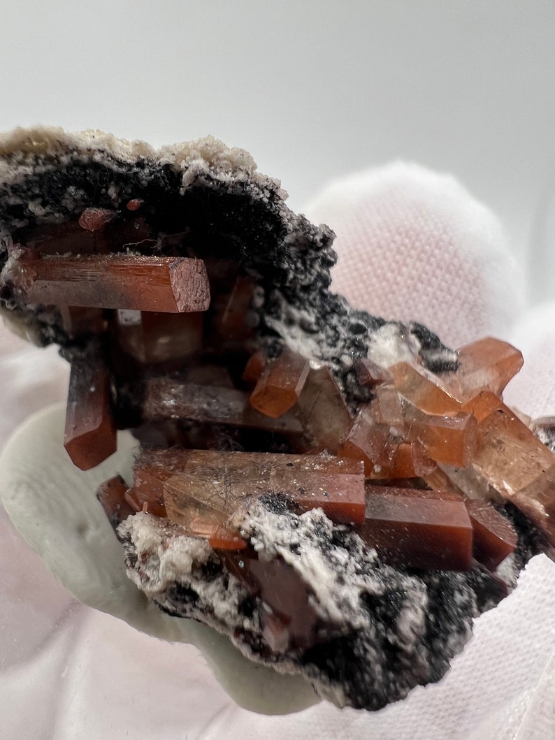 Puede incluir: Primer plano de un esp&eacute;cimen mineral con cristales de color marr&oacute;n rojizo, de forma rectangular. Estos cristales est&aacute;n colocados sobre una base oscura y texturizada con un revestimiento blanco y granulado. Los cristales son transl&uacute;cidos.