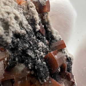 Puede incluir: Primer plano de un esp&eacute;cimen mineral con cristales transl&uacute;cidos de color marr&oacute;n rojizo. Los cristales se agrupan sobre una matriz gris oscuro y blanca. La formaci&oacute;n mineral es texturizada, mostrando la belleza natural del esp&eacute;cimen.