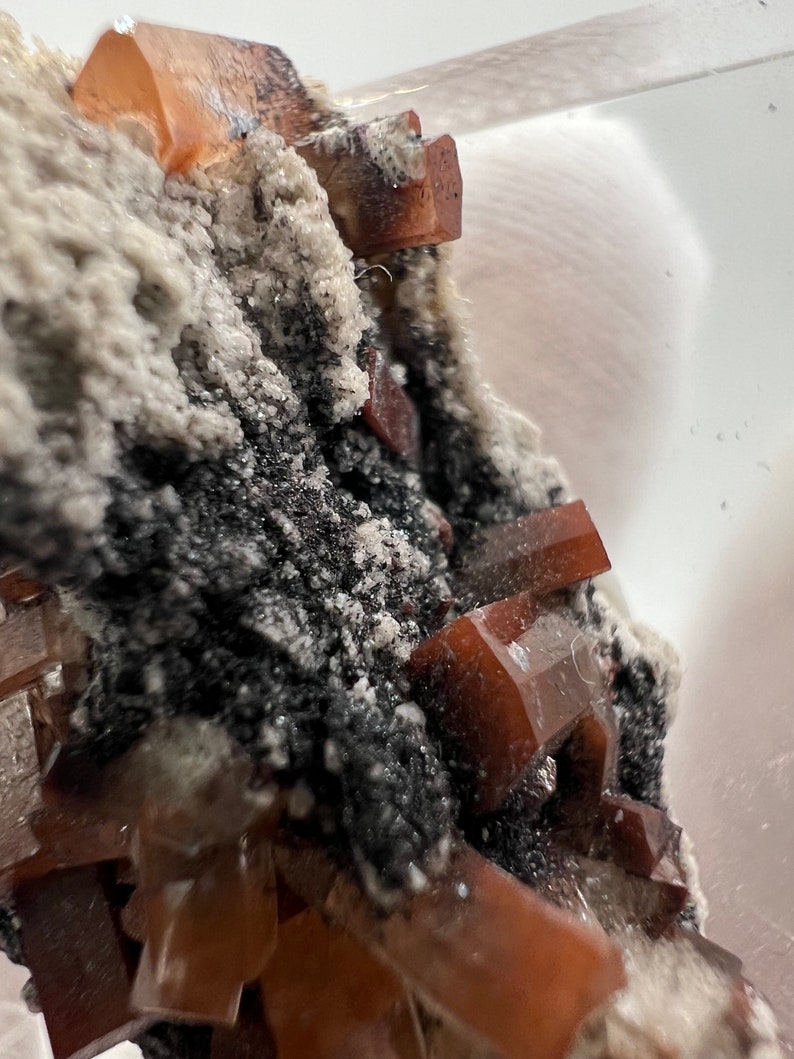 Puede incluir: Primer plano de un esp&eacute;cimen mineral con cristales transl&uacute;cidos de color marr&oacute;n rojizo. Los cristales se agrupan sobre una matriz gris oscuro y blanca. La formaci&oacute;n mineral es texturizada, mostrando la belleza natural del esp&eacute;cimen.