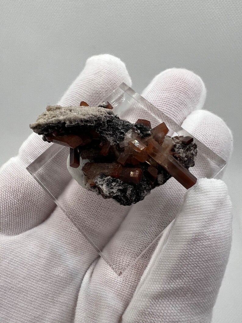 Puede incluir: Un esp&eacute;cimen mineral con cristales de color marr&oacute;n-anaranjado, posiblemente mimetita, presentado en una pantalla acr&iacute;lica transparente. Los cristales est&aacute;n dispuestos sobre una base oscura con zonas blancas y grises.