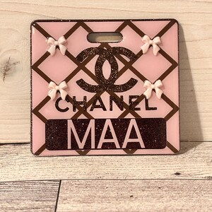 May include: Pink badge holder with a brown glitter design and the word "CHANEL" and the letters "MAA". The badge holder has a brown diamond pattern and four small pink bows.