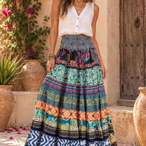 May include: A sleeveless white button-down top paired with a floor-length, high-waisted maxi skirt featuring a smocked waistband and horizontal tiers of multicolored fabric with diverse geometric, floral, and abstract patterns in blue, orange, and green.