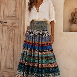 May include: A long, floor-length skirt featuring a wide, smocked elastic waistband and a series of horizontal panels with diverse, colorful patterns in shades of blue, teal, orange, and black, paired with a long-sleeved, button-down white shirt.