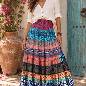 May include: A long, tiered maxi skirt featuring a vibrant patchwork of colorful patterns, including floral, geometric, and ikat-style prints in shades of blue, pink, orange, and black, paired with a long-sleeved, button-down cream-colored blouse.