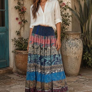 May include: A long, tiered maxi skirt featuring a wide, dark blue elasticized waistband and multiple horizontal panels with diverse, colorful patterns in shades of blue, red, and cream. The skirt is paired with a long-sleeved, button-down cream shirt.