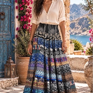 May include: A long, tiered maxi skirt featuring a vibrant patchwork of blue, black, white, and yellow patterns, paired with a cream-colored, long-sleeved button-down shirt with a V-neckline and rolled-up sleeves, worn with simple brown sandals.
