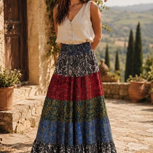 May include: A sleeveless beige V-neck button-down top paired with a long, tiered maxi skirt featuring a vibrant, multi-colored paisley print in shades of black, red, green, and blue, finished with a ruffled hem and brown strappy sandals.