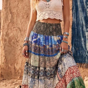 May include: A woman wearing a cream-colored crochet crop top and a long, patchwork skirt. The skirt features various patterns in blue, red, green, and other colors. She is also wearing multiple silver bracelets, rings, and a turquoise necklace.