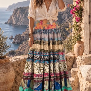 May include: A long, tiered maxi skirt featuring a vibrant patchwork of horizontal bands in various colors and patterns, including floral, geometric, and abstract designs, paired with a light-colored, button-down shirt tied at the waist.