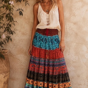 May include: A sleeveless cream-colored top with a V-neckline paired with a long, tiered maxi skirt featuring a wide, smocked waistband and a vibrant, multi-colored pattern of floral, geometric, and abstract prints in shades of blue, red, and orange.