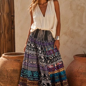 May include: A sleeveless cream-colored V-neck top paired with a floor-length maxi skirt featuring a wide, dark elasticized waistband and a multi-colored patchwork pattern of floral, geometric, and abstract prints in shades of blue, black, and red.