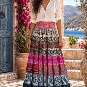 May include: A long, tiered maxi skirt featuring a vibrant patchwork of horizontal bands in various colors and patterns, including floral, geometric, and animal prints, paired with a white, long-sleeved, button-down shirt with rolled-up sleeves.