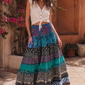 May include: A white sleeveless button-down top with a front tie and a long, tiered maxi skirt featuring a vibrant patchwork of blue, black, and teal floral and geometric patterns with a smocked elastic waistband and brown strappy sandals.