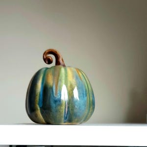 May include: A ceramic pumpkin figurine featuring a glossy glaze with blended shades of teal, blue, and yellow. The pumpkin has a rounded, ribbed body and a prominent, curved brown stem at the top, showcasing a smooth, reflective surface finish.