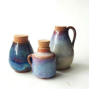 Peut inclure: Trois bouteilles en céramique avec bouchons en liège. Les bouteilles ont une finition brillante avec des teintes bleues, violettes et marron. La bouteille de droite a une poignée. Les bouteilles sont de tailles et de formes différentes.