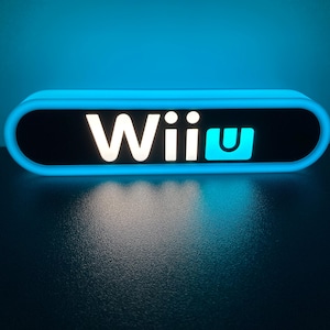 Puede incluir: Un letrero rectangular iluminado con bordes redondeados, que muestra el texto "Wii U" en blanco y turquesa sobre un fondo negro. El letrero está rodeado por una luz azul brillante, lo que sugiere un tema de juego.