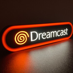 May include: A black and orange neon sign with the text "Dreamcast" and a stylized orange swirl logo.