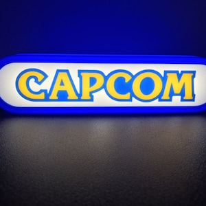 May include: A blue and white oval sign with the word "CAPCOM" in yellow letters.