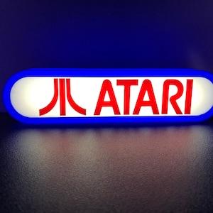 May include: A blue and white oval sign with the red text "ATARI" and the Atari logo. The sign is illuminated from behind.