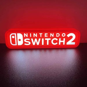 May include: A red rectangular sign with the words "NINTENDO SWITCH 2" in white lettering. The sign also features the Nintendo Switch logo. The sign is illuminated and sits on a dark surface.