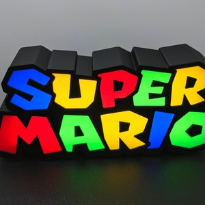 May include: A black light-up sign with the words "SUPER MARIO" in colorful 3D letters. The letters are blue, yellow, green, and red.