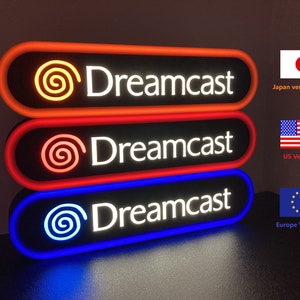 May include: Three Dreamcast logo signs with different colored borders. The top sign has an orange border, the middle sign has a red border, and the bottom sign has a blue border. Each sign has the Dreamcast logo and the text "Dreamcast" in white.