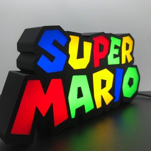 May include: A black light-up sign with the words "SUPER MARIO" in colorful, 3D letters. The letters are outlined in black and have a textured surface. The sign is lit up with different colors, including red, green, blue, and yellow.