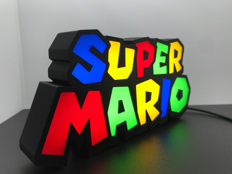 May include: A black light-up sign with the words "SUPER MARIO" in colorful, 3D letters. The letters are outlined in black and have a textured surface. The sign is lit up with different colors, including red, green, blue, and yellow.