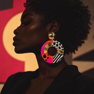 Puede incluir: Un pendiente circular grande con un poste dorado y un aro multicolor. El aro muestra segmentos en rosa neón, naranja brillante, estampados de animales en blanco y negro, y detalles de metal dorado pulido en un diseño geométrico llamativo.