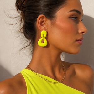 May include: A woman wearing bright yellow earrings and a gold chain necklace. The earrings have a geometric design. She is also wearing a neon yellow one-shoulder top.