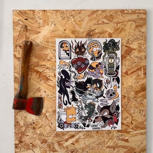 Op de afbeelding: Een bijl met een houten handvat en een rood blad naast een vel stickers met diverse illustraties in cartoonstijl. De sticker vellen bevatten afbeeldingen van karakters en objecten in tattoo-stijl.