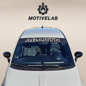 Könnte beinhalten: Ein weißer Fiat 500 Abarth mit einem schwarzen Banner über der Windschutzscheibe mit der weißen Aufschrift "ABARTH". Das Auto hat schwarze Seitenspiegel und ein schwarzes Emblem vorne.
