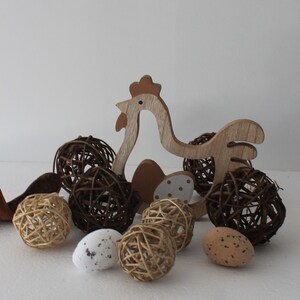 Peut inclure: Décorations sur le thème de Pâques avec des découpes de poulets en bois, des sphères tressées et des œufs décoratifs. Les œufs présentent des couleurs et des motifs variés, notamment des motifs mouchetés et à pois. L'ensemble est posé sur un fond blanc.