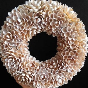 Op de afbeelding: Een krans gemaakt van natuurlijke beige pistache-schalen, gerangschikt in een cirkelvorm om op bloemen te lijken. De krans heeft een diameter van ongeveer 30 cm.