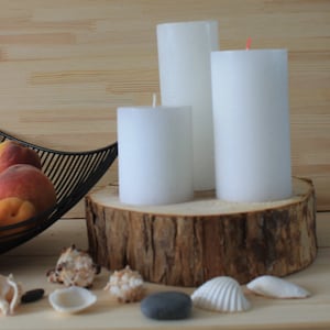 May include: Three white pillar candles of varying heights are arranged on a wooden log slice. The candles are on a wooden surface with seashells and stones scattered around them.
