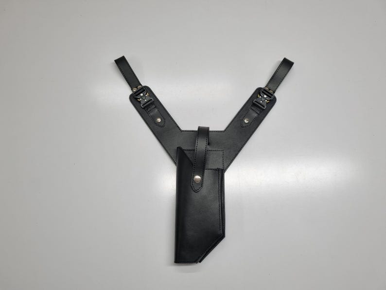 May include: A black leather shoulder holster featuring a Y-shaped harness design with two adjustable straps, metal buckles, and a central vertical pouch secured by a single snap button closure. The construction includes reinforced stitching and rivets.