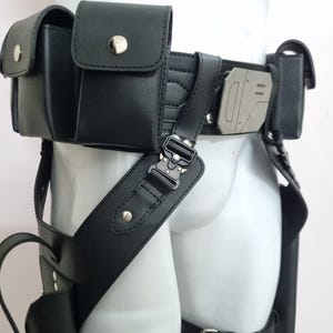 May include: A black utility belt featuring multiple pouches with silver-toned snap closures, a metallic geometric buckle detail, and adjustable straps with tactical-style hardware. The belt is constructed from a smooth material and includes leg holsters.