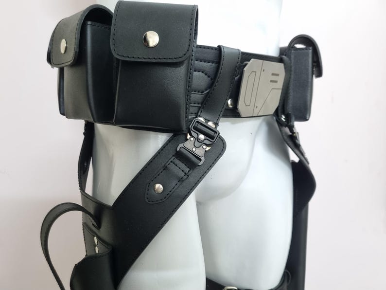 May include: A black utility belt featuring multiple pouches with silver-toned snap closures, a metallic geometric buckle detail, and adjustable straps with tactical-style hardware. The belt is constructed from a smooth material and includes leg holsters.
