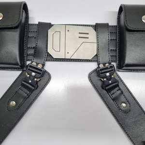 May include: A black utility belt featuring two rectangular pouches with silver snap buttons, a central metallic plate with engraved geometric lines, and two adjustable straps connected by black metal quick-release buckles with silver rivets.
