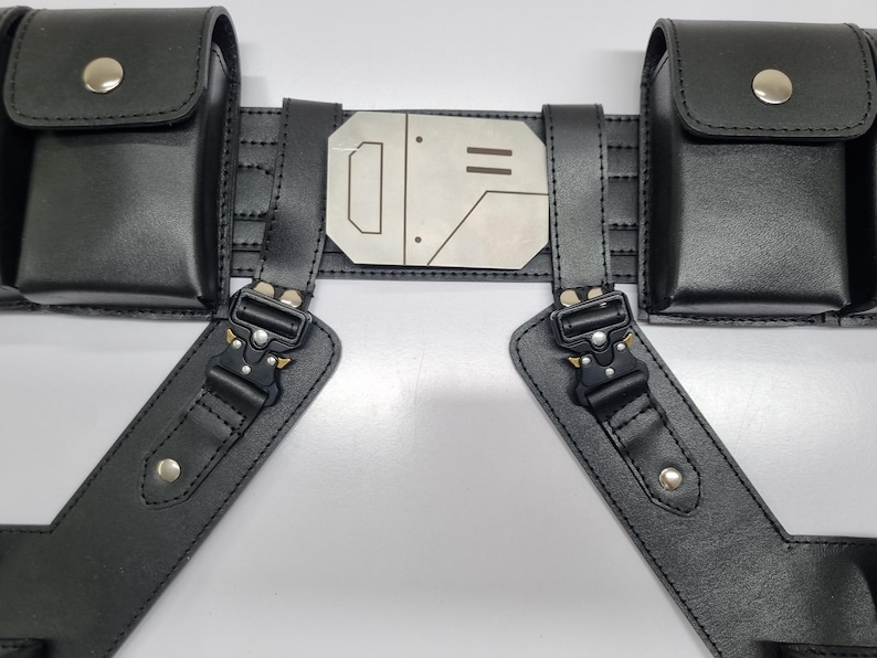 May include: A black utility belt featuring two rectangular pouches with silver snap buttons, a central metallic plate with engraved geometric lines, and two adjustable straps connected by black metal quick-release buckles with silver rivets.
