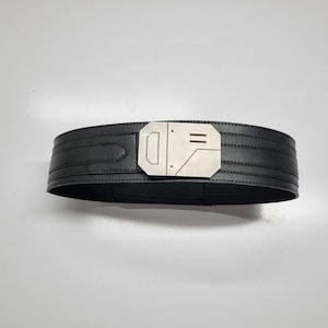 May include: A black belt featuring a wide, structured band with horizontal stitched detailing and a prominent, geometric silver-toned metal buckle. The buckle has a flat, angular design with engraved lines and small circular accents on its surface.