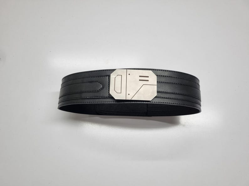 May include: A black belt featuring a wide, structured band with horizontal stitched detailing and a prominent, geometric silver-toned metal buckle. The buckle has a flat, angular design with engraved lines and small circular accents on its surface.