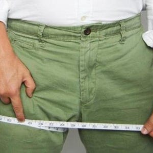 May include: Olive green trousers featuring a button fly and front pockets, shown with a white measuring tape wrapped around the thigh area, indicating a measurement of approximately 25 inches across the upper leg section of the garment.
