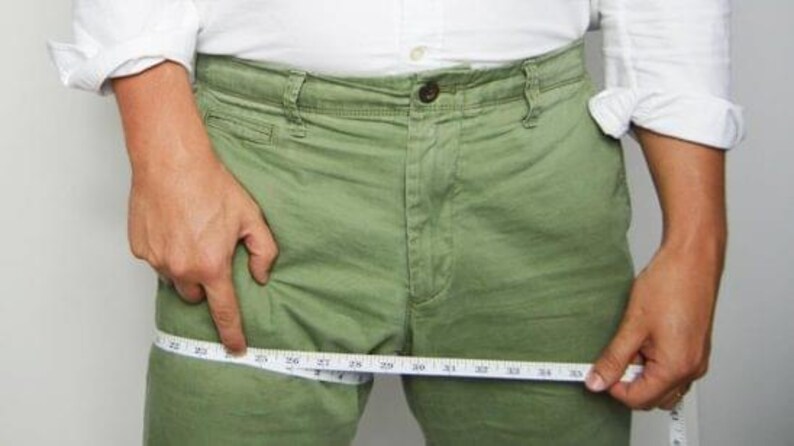 May include: Olive green trousers featuring a button fly and front pockets, shown with a white measuring tape wrapped around the thigh area, indicating a measurement of approximately 25 inches across the upper leg section of the garment.