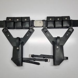 May include: A black utility belt featuring six rectangular pouches with silver snap closures, a central metallic buckle, and two integrated holsters suspended from Y-shaped straps. The assembly includes adjustable leg straps with silver buckles.